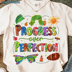 May include: White t-shirt with a colorful graphic design featuring a green caterpillar, a butterfly, and the words "PROGRESS over PERFECTION." The design includes an orange sun, apples, and a dragonfly.