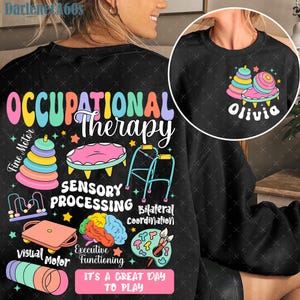 Puede incluir: Sudadera negra con las palabras "OCCUPATIONAL THERAPY" en letras coloridas. El diseño incluye imágenes de herramientas de terapia, un cerebro y el texto "IT'S A GREAT DAY TO PLAY". Un diseño más pequeño a la derecha dice "Olivia".