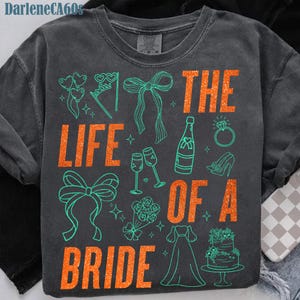 Może przedstawiać: Ciemnoszara bluza z napisem "THE LIFE OF A BRIDE" w pomarańczowym brokatowym napisie. Projekt zawiera turkusowe ilustracje kokard, szampana, pierścionka, sukni i ciasta.