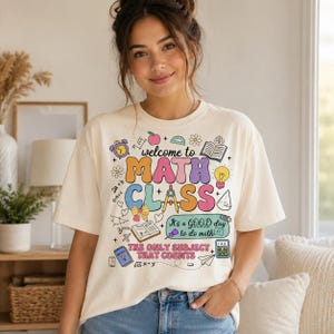 May include: A cream-colored t-shirt featuring a colorful graphic with the text "welcome to MATH CLASS", "It's a GOOD day to do math", and "THE ONLY SUBJECT THAT COUNTS". The design includes illustrations of an apple, clock, book, lightbulb, and calculator.