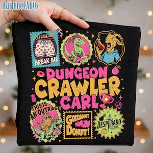 Puede incluir: Camiseta negra con gráficos coloridos y texto. El diseño incluye las palabras "DUNGEON CRAWLER CARL" y otras frases como "YOU WILL NOT BREAK ME" y "GODDAMN DONUT!". La camiseta presenta varias ilustraciones de dibujos animados.