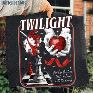 Puede incluir: Camiseta negra con la palabra "TWILIGHT" en blanco. El diseño incluye una manzana roja sostenida en manos, una cinta roja y piezas de ajedrez. También está presente el texto "...And so the lion fell in love with the lamb".