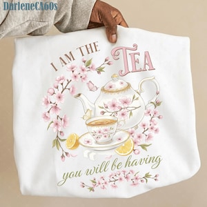 Può includere: Canovaccio bianco con un design floreale per un tea party. Il design include una teiera, una tazza da tè e fette di limone, circondate da fiori di ciliegio rosa e farfalle. Il testo recita "I AM THE TEA you will be having."