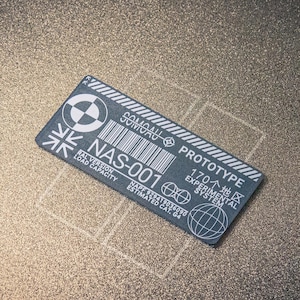 Könnte beinhalten: Ein rechteckiges, graues NAS-001 Prototyp-Etikett mit weißem Text und einem Barcode, das auf einer strukturierten, hellbraunen Oberfläche platziert ist. Das Etikett enthält ein kreisförmiges Logo, eine Flagge und den Text "170 Experimental System".