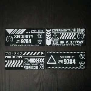 Könnte beinhalten: Vier schwarz-weiße Aufkleber mit futuristischem Design. Die Aufkleber zeigen den Text "SECURITY UNIT 9784" und "EXPERIMENTAL 実験的 CAUTION 注意 RK V. 3.11". Ein Aufkleber enthält auch den Text "PROTOTYPE" und "SERIAL/29482 ARC X9".