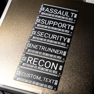 Pode incluir: Etiquetas retangulares pretas com texto branco, incluindo "ASSAULT", "SUPPORT", "SECURITY", "NETRUNNER", "RECON" e "CUSTOM_TEXT". Cada etiqueta tem um padrão único e um design semelhante a um código de barras. As etiquetas estão em uma superfície metálica texturizada.
