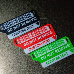 Puede incluir: Tres etiquetas rectangulares con diseño de código de barras. Las etiquetas son negras, rojas y verdes, cada una con el texto "DO NOT REMOVE!" y "CUSTOM TEXT" en blanco. Las etiquetas tienen un pequeño agujero en un extremo.
