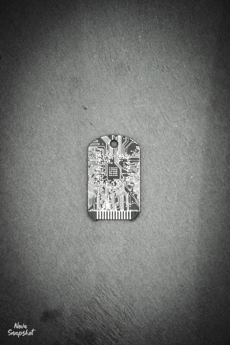 Op de afbeelding: Een zilverkleurig metalen tag met een printplaatontwerp. De tag is rechthoekig met afgeronde hoeken en heeft een klein gaatje bovenaan.
