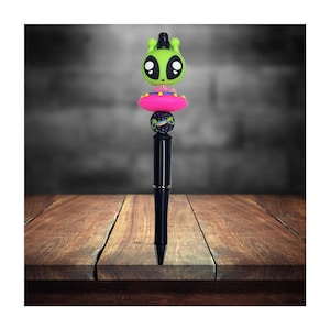 May include: A black pen with a green alien figurine on top. The alien is wearing a pink flying saucer hat and has big eyes. The pen is on a wooden surface.