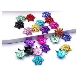 May include: A collection of star-shaped beads in various colors, including blue, green, red, pink, and gold. Each bead has a central circular design and is scattered on a white surface. These beads are likely used for jewelry making or craft projects.