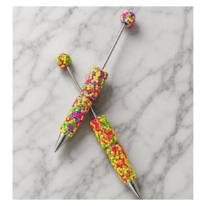 May include: Two pens featuring cylindrical barrels and spherical toppers covered in a textured, multi-colored sprinkle pattern of neon pink, yellow, green, and blue dots. The pens have silver-toned metal tips and thin, straight metal shafts.