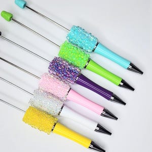 May include: A collection of colorful pens with a beaded grip and a silver metal shaft. The pens feature a variety of colors, including blue, green, purple, pink, white, and yellow. The pen tips are black.