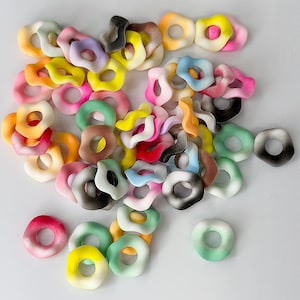 May include: A collection of colorful, abstract donut-shaped polymer clay beads in various sizes and shades. The beads feature a mix of colors, including pink, yellow, green, orange, and black, with some having a gradient effect. These beads are ideal for jewelry making.