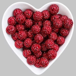 May include: A collection of round, deep red beads with a textured, sparkling surface finish, contained within a white, heart-shaped bowl. Each bead features a small central hole, and the vibrant red color is consistent across all the pieces.