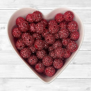 May include: A heart-shaped white bowl filled with numerous red, sparkly beads. The beads have a textured surface and are a deep, rich red color. The bowl sits on a white, wooden surface.