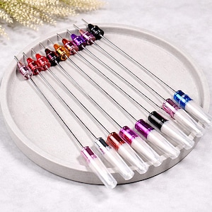 May include: A collection of lip gloss applicators with various colored caps, including red, pink, purple, and black, arranged on a gray, circular tray. Each applicator has a clear tube and a thin, silver wand.
