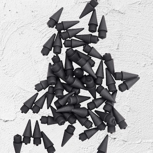 May include: A collection of dark gray pencil tips with a conical shape and threaded base, scattered on a textured white surface. The tips are uniformly colored and appear to be made of a matte material.