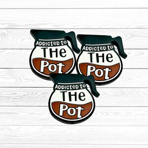 May include: Three enamel pins shaped like coffee carafes with black outlines and handles. The white center features the black text ADDICTED TO THE POT, with a brown section at the bottom representing coffee. The design is consistent across all three pins.