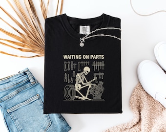May include: Black t-shirt with a graphic of a skeleton working on an engine, with the text "WAITING ON PARTS". The design includes tools and a tire. A silver necklace is draped across the shirt.
