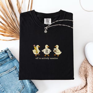 May include: Black t-shirt featuring a graphic of three cartoon ducks, each with a pencil, a sign saying "You Got This", and a coffee cup. The text "off to actively monitor" is below. A silver necklace rests on the shirt.