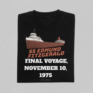 May include: Black t-shirt featuring a brown and white illustration of the SS Edmund Fitzgerald, a freighter ship. The text reads "SS Edmund Fitzgerald, Final Voyage, November 10, 1975" in orange and white. The shirt is folded.