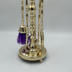Puede incluir: Un juego de herramientas para chimenea de oro con cepillo, pala, atizador y pinzas. Las herramientas están montadas en un soporte decorativo con una base redonda y tres patas.