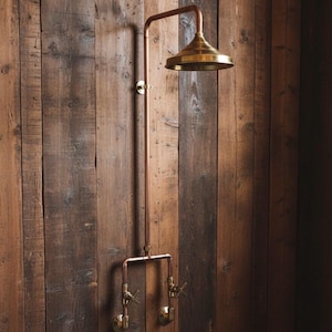 May include: A copper and brass shower fixture, mounted on a wooden wall. The showerhead is a round, brass design. The fixture includes two brass faucet handles. The copper pipes are visible.