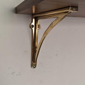May include: A brass shelf bracket is mounted on a white wall, supporting a wooden shelf. The bracket has a decorative, curved design and is attached with screws. The shelf is a warm brown color.