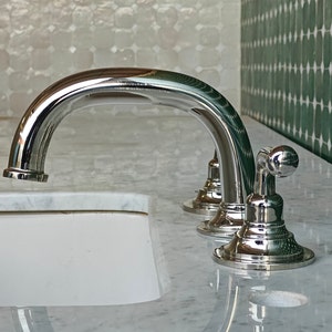 Puede incluir: Un grifo de baño cromado con un caño curvo y dos manijas. El grifo está montado en una encimera de mármol blanco.