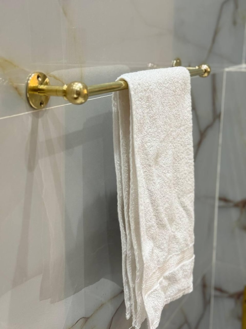 Styled vignette: warm raw brass towel bar against stone tile with folded linens and greenery. Perfect upgrade for guest bath, Airbnb, or modern farmhouse interiors; mixes well with marble and wood accents.