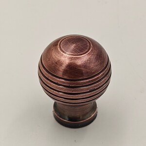 Puede incluir: Un pomo de armario redondo de color cobre con un dise&ntilde;o texturizado y acanalado. El pomo tiene una parte superior circular ligeramente elevada.