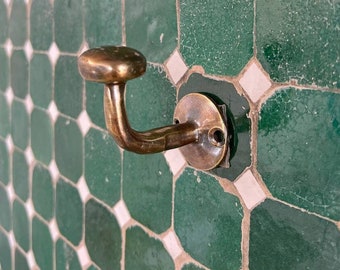 May include: A brass hook with a rounded top and a curved arm, mounted on a green and white tiled wall.