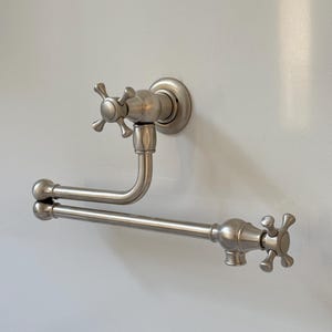 May include: A brushed nickel wall-mounted pot filler faucet with cross-shaped handles. The faucet has a curved spout and a horizontal bar for holding a towel or other items. The fixture is mounted on a white wall.
