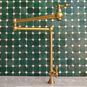 May include: A gold-coloured, articulated pot filler tap against a backdrop of green square tiles. The tap has a vertical base, a horizontal arm, and a swivelling spout. The design is reminiscent of a professional kitchen.