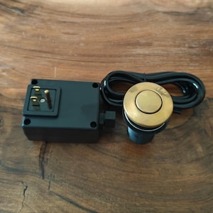 May include: A black electrical component with a three-prong plug, a brass-colored circular button, and a black cord. The components are arranged on a wooden surface.