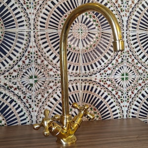 May include: A gold-colored faucet with cross-shaped handles and a curved spout. The faucet is set against a backdrop of patterned tiles in shades of blue, green, and brown. The faucet is on a wooden surface.