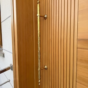 May include: A vertical, gold-coloured door pull with ornate detailing is mounted on a light brown wooden door. The door has vertical, grooved panels. The pull is attached with two circular, gold-coloured mounts.