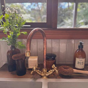 May include: A copper faucet with gold-colored handles is the focal point of this kitchen scene. A brown glass bottle with a plant, a soap bar, and a soap dispenser are on the counter. A window with a view of trees is in the background.