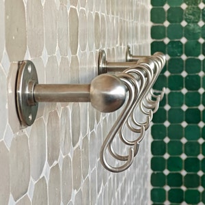 Puede incluir: Un perchero de pared de acero inoxidable con múltiples ganchos curvos. El perchero está montado en una pared con un diseño de mosaico en tonos grises y verdes. Los ganchos están diseñados con un estilo decorativo y fluido.