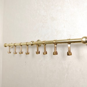 May include: A brass coat rack with eight hooks mounted on a white wall. The rack has a round end cap and is made of solid brass.