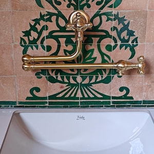 Puede incluir: Un lavabo de porcelana blanca con un grifo y una barra para toallas de color dorado. El lavabo tiene un desagüe con una tapa dorada. El lavabo está colocado contra una pared con azulejos de patrones verdes y marrones. El lavabo está etiquetado como "Vida".