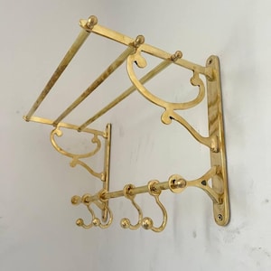 May include: A vintage brass wall-mounted coat rack with a shelf and multiple hooks. The rack features ornate, curved designs and is mounted on a white wall. The brass has a warm, golden hue, adding a touch of classic elegance.