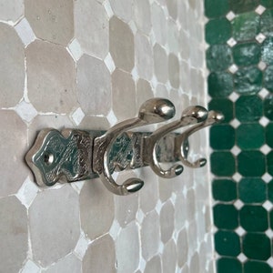 May include: Silver metal coat rack with three double hooks. The rack has an ornate design and is mounted on a white tiled wall.