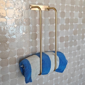 Unlacquered brass towel rack wall-mounted in a spa-style bathroom with white subway tile; solid raw brass bar holds plush towels and doubles as bathroom décor and towel organizer; living-finish patina-ready surface adds antique charm to modern or vintage bath remodels.