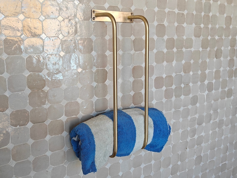 Unlacquered brass towel rack wall-mounted in a spa-style bathroom with white subway tile; solid raw brass bar holds plush towels and doubles as bathroom décor and towel organizer; living-finish patina-ready surface adds antique charm to modern or vintage bath remodels.