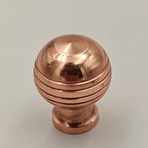 Puede incluir: Un pomo de armario de cobre con una parte superior redonda y acanalada y una base lisa y redondeada.