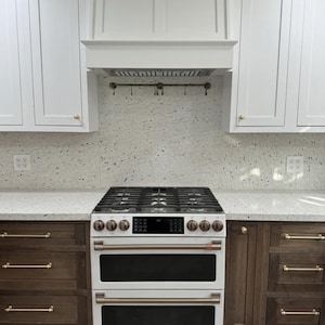 Pode incluir: Uma cozinha moderna com um fogão a gás em aço inoxidável e um forno duplo. Armários brancos com puxadores dourados estão acima dos armários inferiores castanhos escuros. O backsplash é salpicado. Uma exaustor branco está acima do fogão.