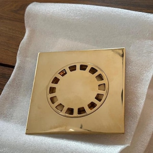 Puede incluir: Una tapa de desagüe cuadrada de color dorado con un patrón circular de aberturas rectangulares en el centro. La tapa está hecha de un metal brillante y reflectante y se encuentra sobre una superficie blanca texturizada.