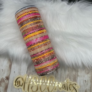 May include: A stainless steel tumbler with a colorful, striped design featuring pink, yellow, orange, and brown rhinestones. The tumbler is on a white fluffy surface.