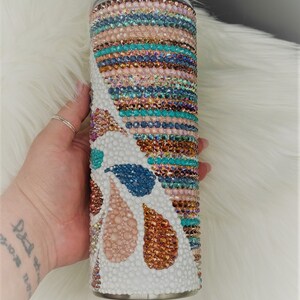 May include: A white tumbler with a colorful design of turquoise, pink, and brown rhinestones. The design features a wave pattern and abstract shapes.